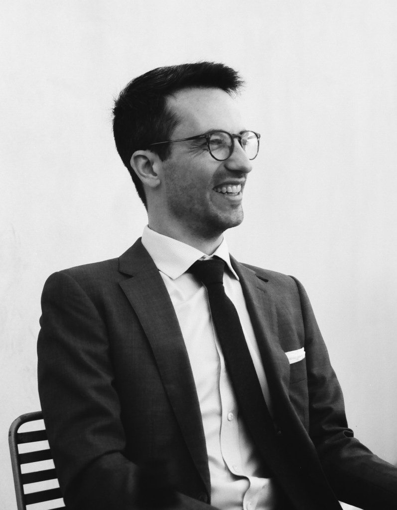Simon Liebling in a black-and-white photograph, seating, wearing a suit